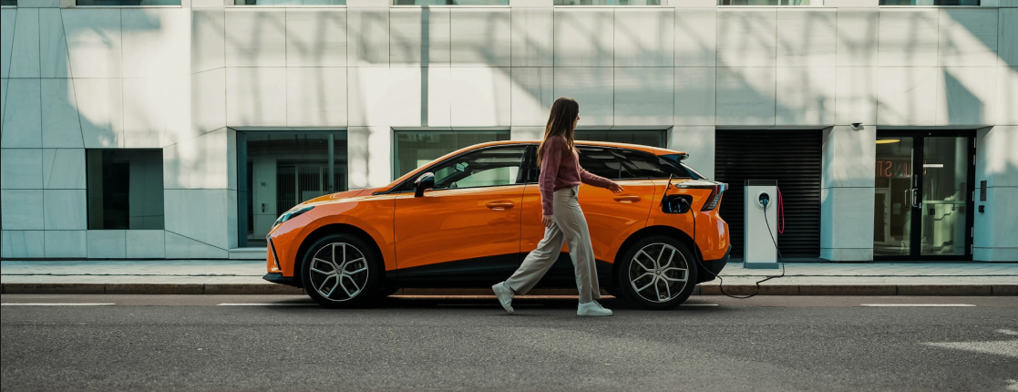 Image of a woman walking in front of a parked orange MG 4 EV on a city road
